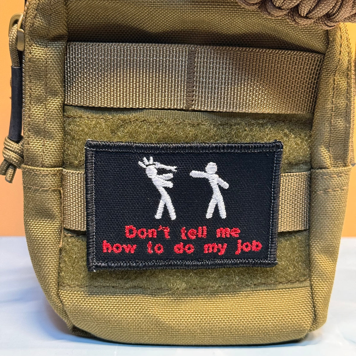 A close-up of a tactical utility pouch in olive drab/khaki nylon with a black rectangular embroidered patch on the front. The patch features two white stick figures and red text that reads 'Don't tell me how to do my job'.