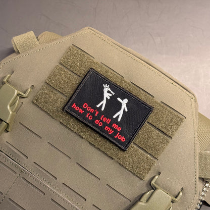 A close-up of a tactical bag in coyote tan with a black rectangular patch on the front. The patch features white stick figures and red text that reads 'Don't tell me how to do my job'.