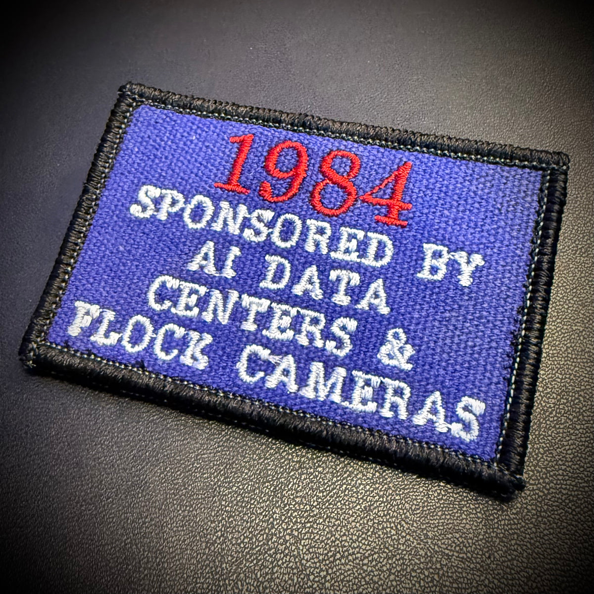 A rectangular embroidered patch with a blue background and black stitched border, featuring the text '1984 SPONSORED BY AI DATA CENTERS & FLOCK CAMERAS' in red and white lettering, placed on a dark textured surface.