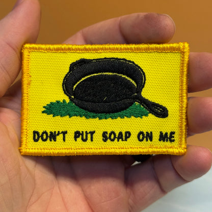 A rectangular embroidered patch held in a hand, featuring a yellow background with a black frying pan sitting on green grass and the text 'DON'T PUT SOAP ON ME' in black.