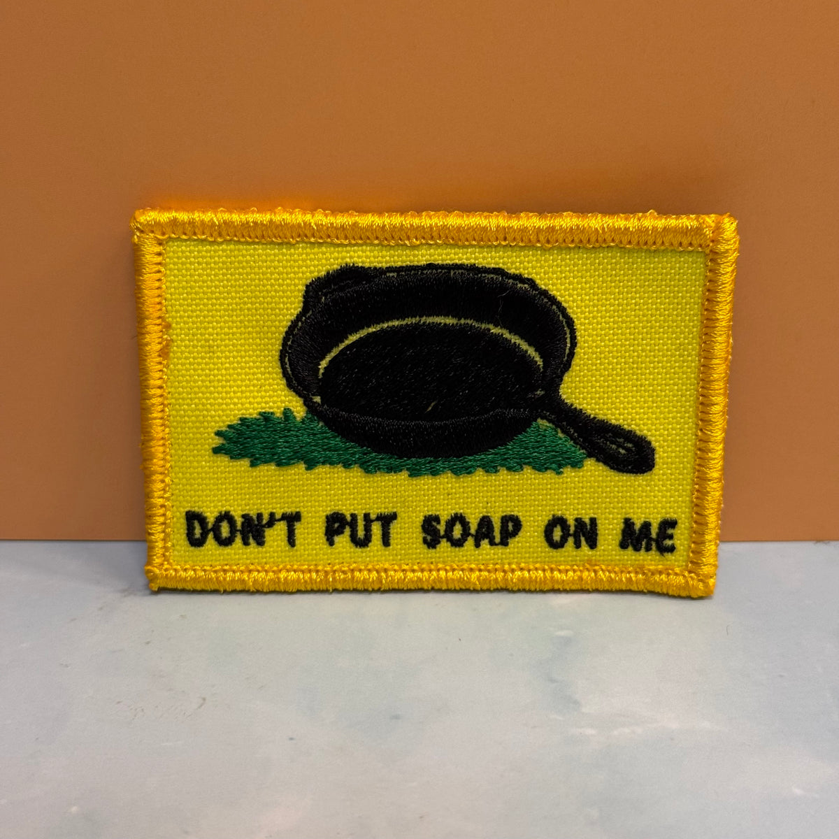 A rectangular embroidered patch with a yellow border and yellow background, featuring a black frying pan above green grass and the text 'DON'T PUT SOAP ON ME' in black. The patch is photographed against a tan wall and a light gray surface.