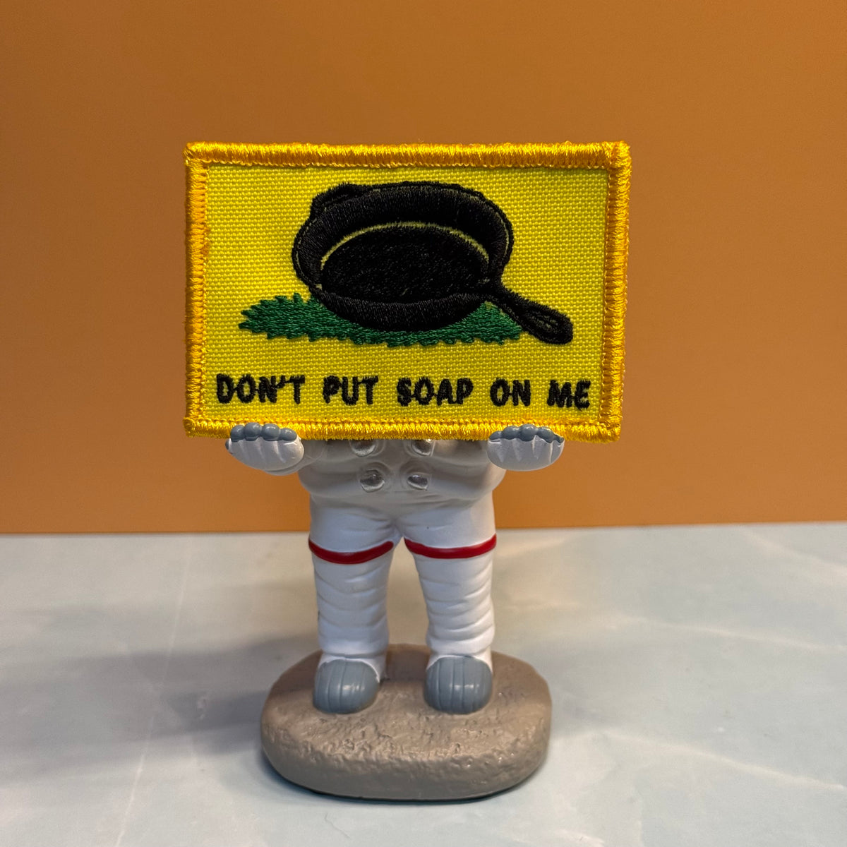 A small embroidered patch with a yellow border and a black frying pan above the text 'DON'T PUT SOAP ON ME' is held by a white astronaut figurine against an orange background.