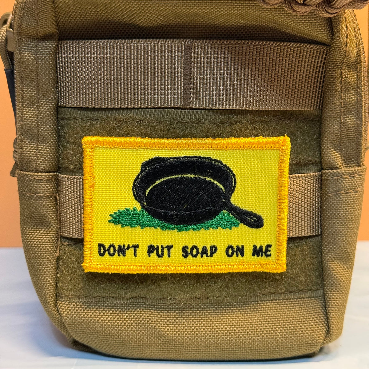 A tan tactical pouch with a yellow embroidered patch featuring a black frying pan and the text 'DON'T PUT SOAP ON ME' on a tan background.