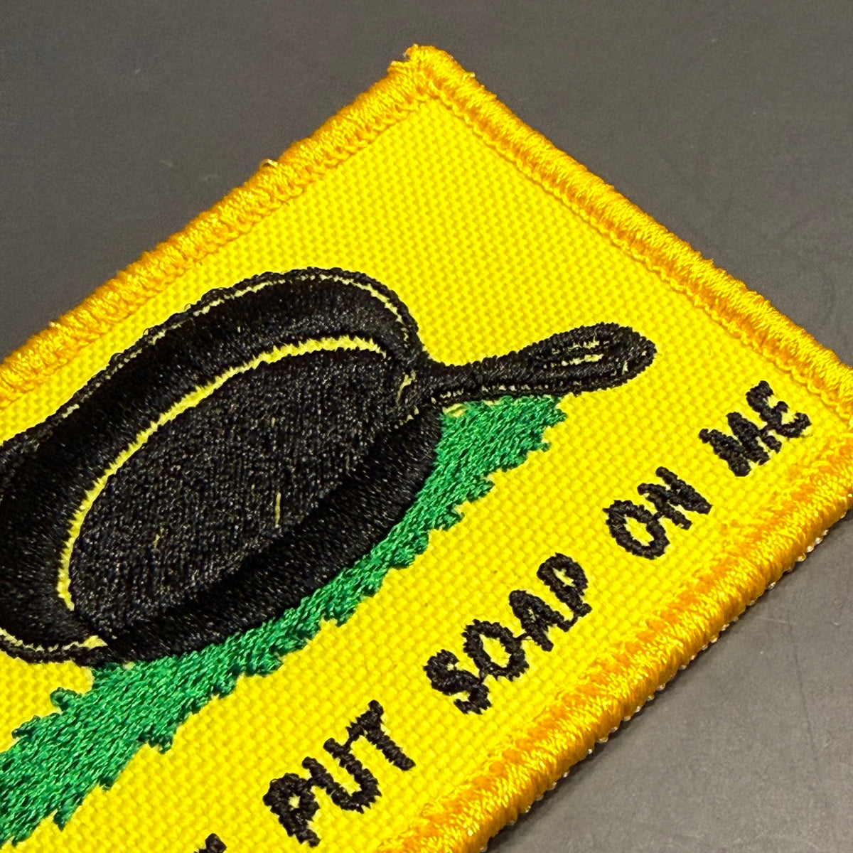 A close-up of a yellow embroidered patch with a black frying pan graphic, green grass-like shape, and black text reading 'PUT SOAP ON ME' on a dark gray background.