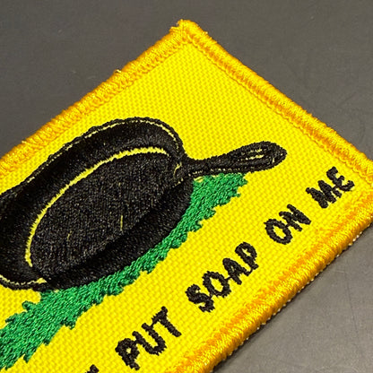 A close-up of a yellow embroidered patch with a black frying pan graphic, green grass-like shape, and black text reading 'PUT SOAP ON ME' on a dark gray background.