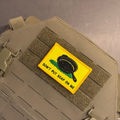 A rectangular tactical patch with a yellow background, featuring a black frying pan on green grass and the text 'DON'T PUT SOAP ON ME', attached to a tan tactical bag with MOLLE webbing.