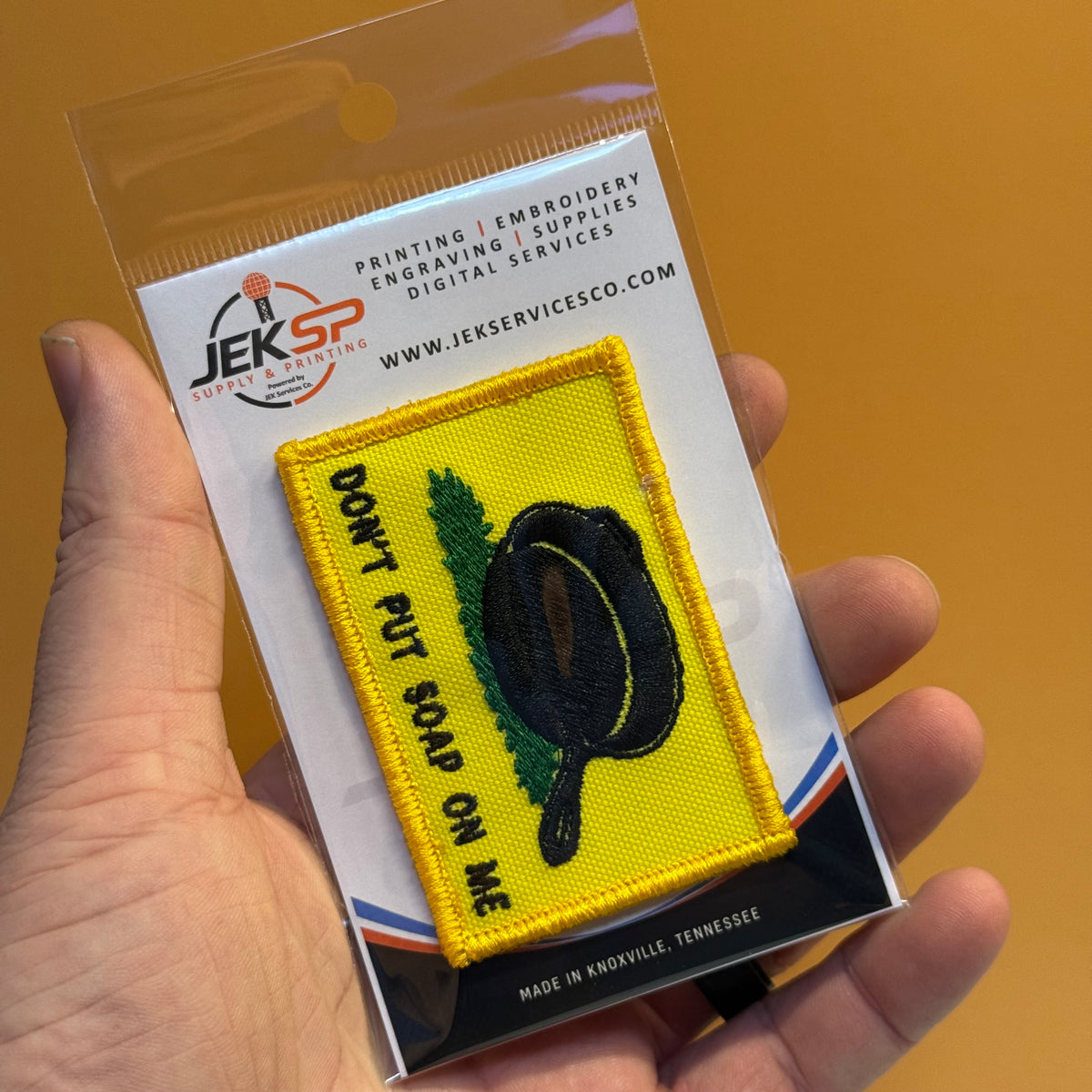A hand holds a small embroidered patch in its retail backing card. The patch is bright yellow with a black pan graphic, green leaves, and the text 'DON'T PUT SOAP ON ME.