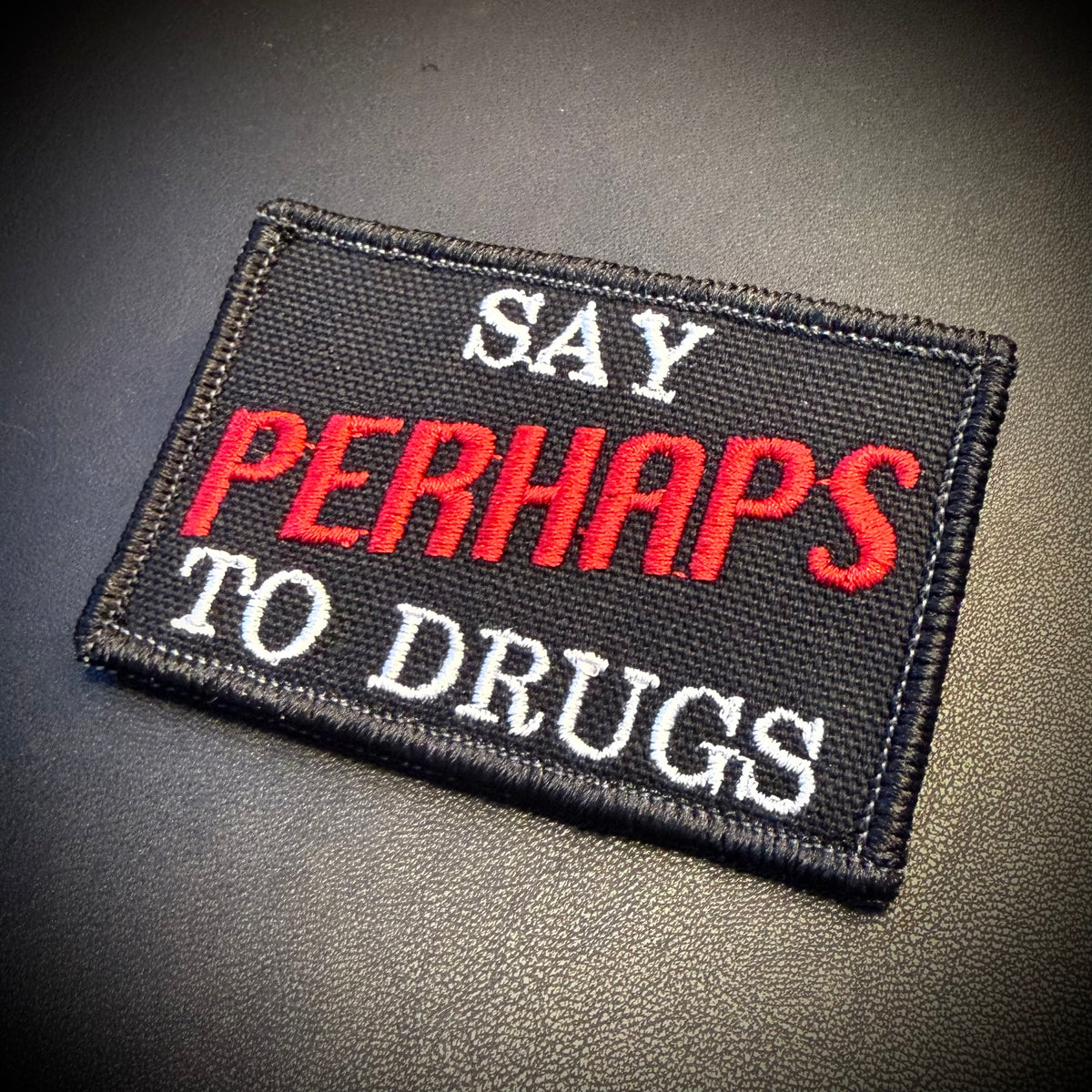 A rectangular embroidered patch with the text 'SAY PERHAPS TO DRUGS' in white and red lettering on a black background, placed on a dark textured surface.