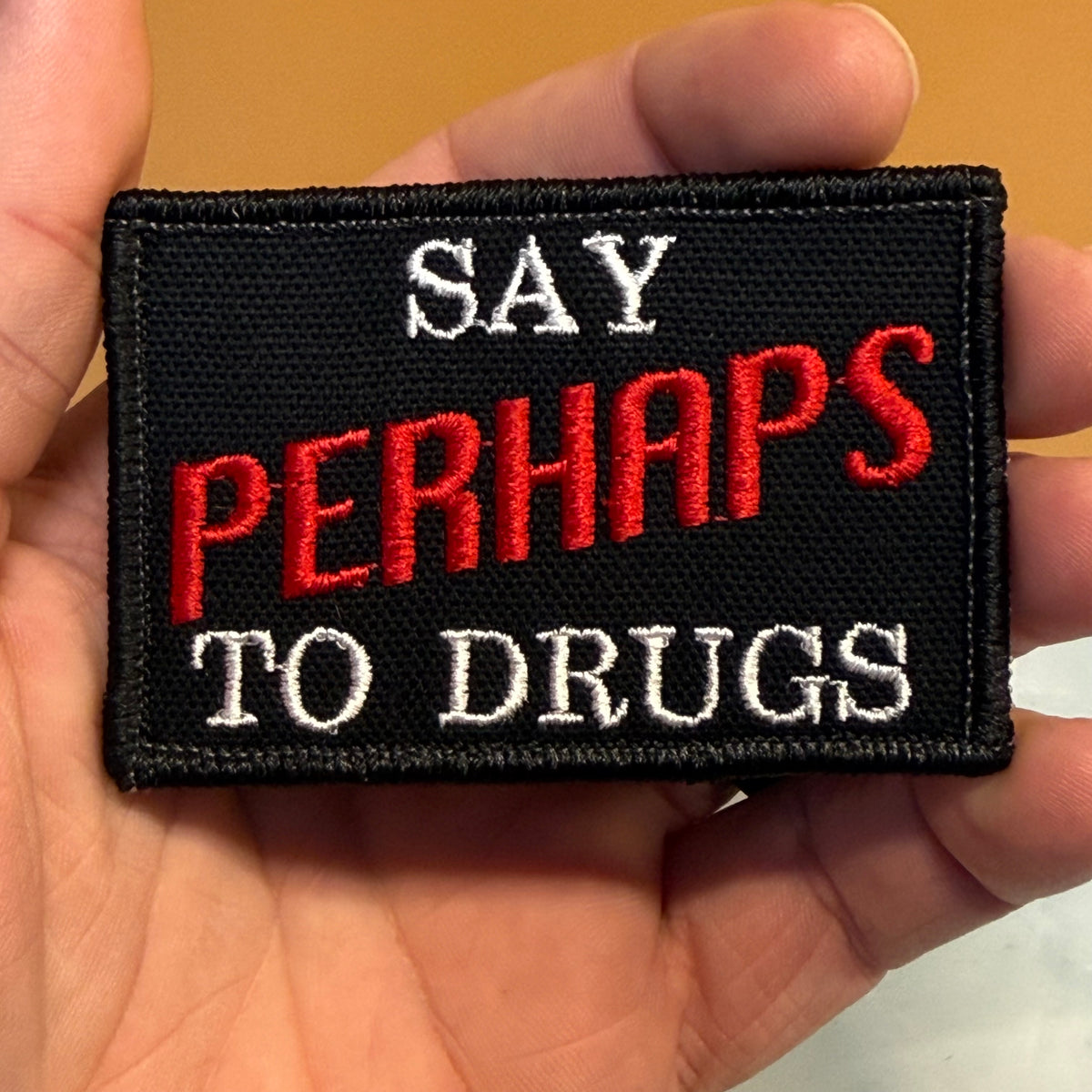 A rectangular black embroidered patch held in a hand, featuring the text 'SAY PERHAPS TO DRUGS' in white and red lettering.