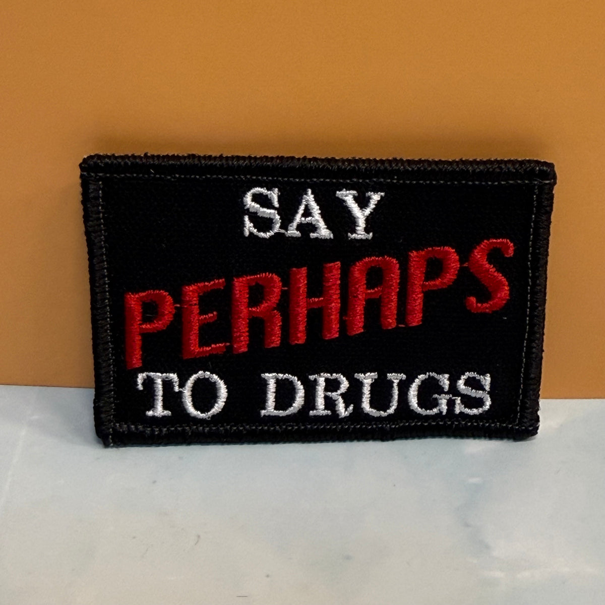 A rectangular embroidered patch with a black background and the text 'SAY PERHAPS TO DRUGS' in white and red lettering, displayed against a warm orange wall and light marble surface.