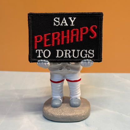 A novelty astronaut figurine holding a black embroidered patch that reads 'SAY PERHAPS TO DRUGS' in white and red lettering, set against an orange background and light marble surface.