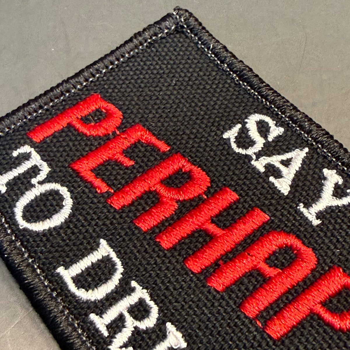 A close-up of a black embroidered patch with red and white text reading 'PERHAPS I'D DRAIN TO DRI' on a gray textured background.