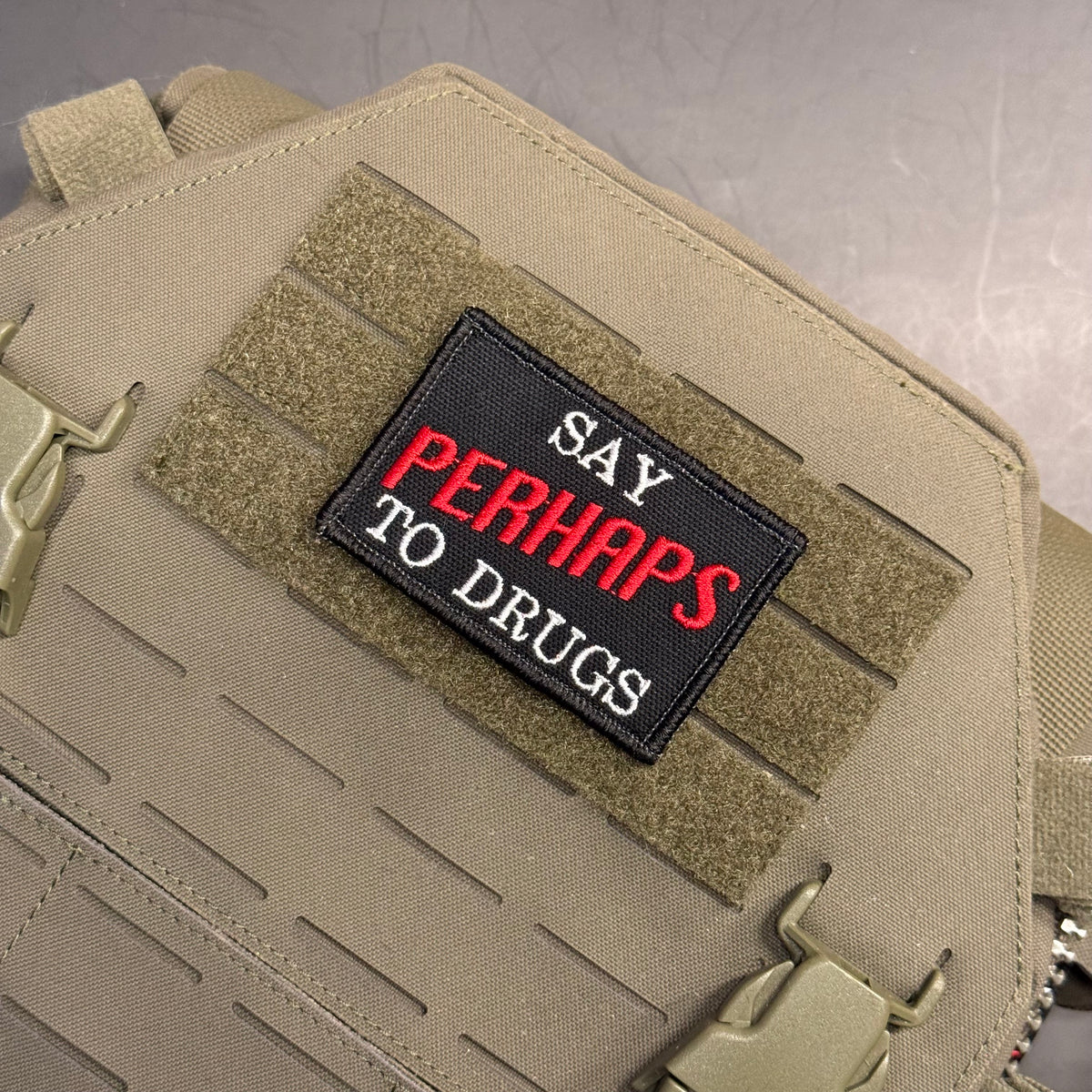 A tactical patch with the text 'SAY PERHAPS TO DRUGS' in white and red lettering on a black background, mounted on tan MOLLE webbing of a tactical vest or bag.