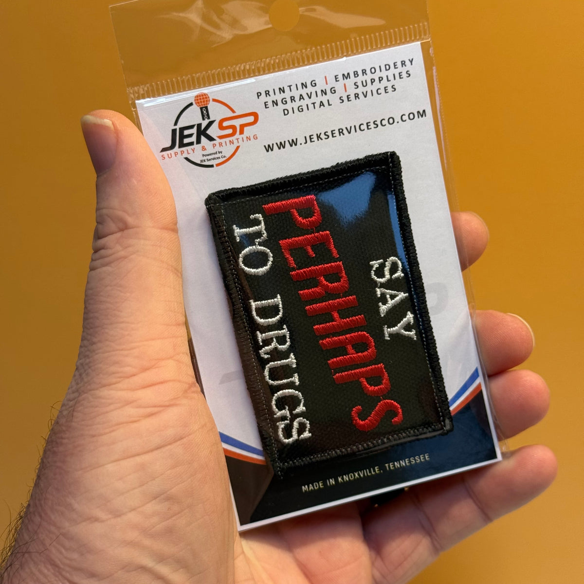 A hand holds a small embroidered patch in its retail packaging against a solid mustard-yellow background. The patch is black with white and red text reading 'SAY PERHAPS TO DRUGS'.