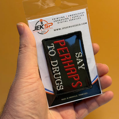 A hand holds a small embroidered patch in its retail packaging against a solid mustard-yellow background. The patch is black with white and red text reading 'SAY PERHAPS TO DRUGS'.
