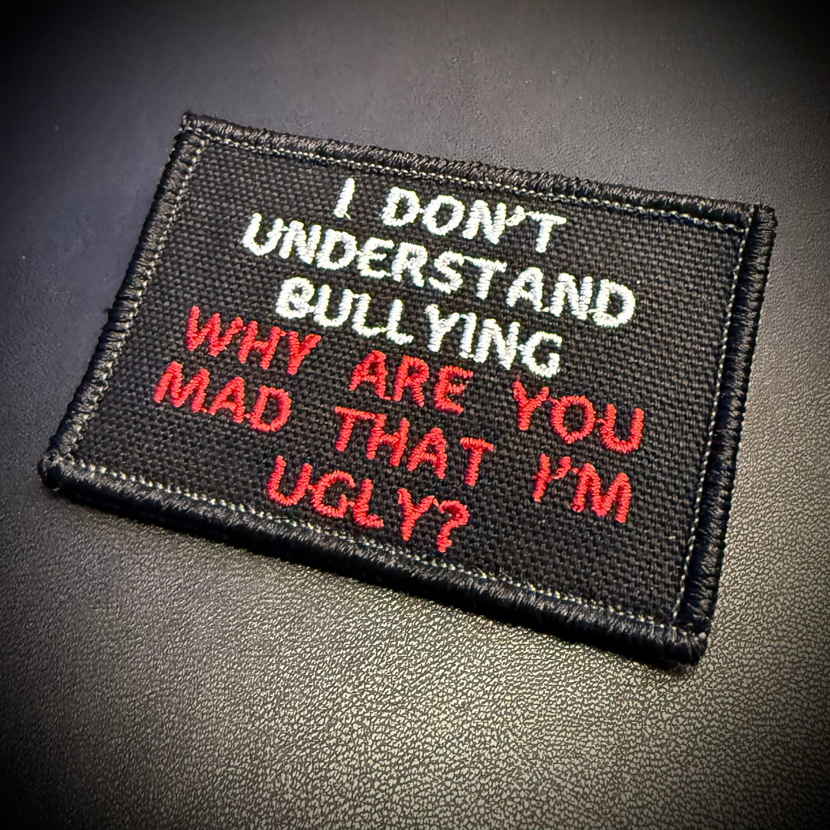 A rectangular black embroidered patch with white and red text reading 'I DON'T UNDERSTAND BULLYING WHY ARE YOU MAD THAT I'M UGLY?' placed on a dark textured surface.