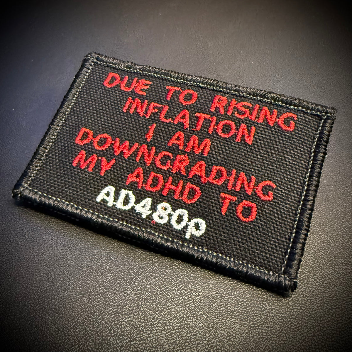 A rectangular black embroidered patch with red and white text reading 'DUE TO RISING INFLATION I AM DOWNGRADING MY ADHD TO AD480P', photographed on a dark textured surface.