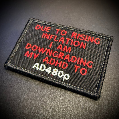 A rectangular black embroidered patch with red and white text reading 'DUE TO RISING INFLATION I AM DOWNGRADING MY ADHD TO AD480P', photographed on a dark textured surface.
