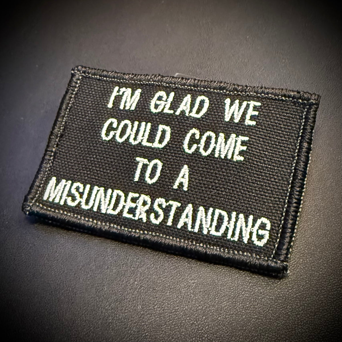 A rectangular black embroidered patch with white text reading 'I'M GLAD WE COULD COME TO A MISUNDERSTANDING' on a dark textured background.