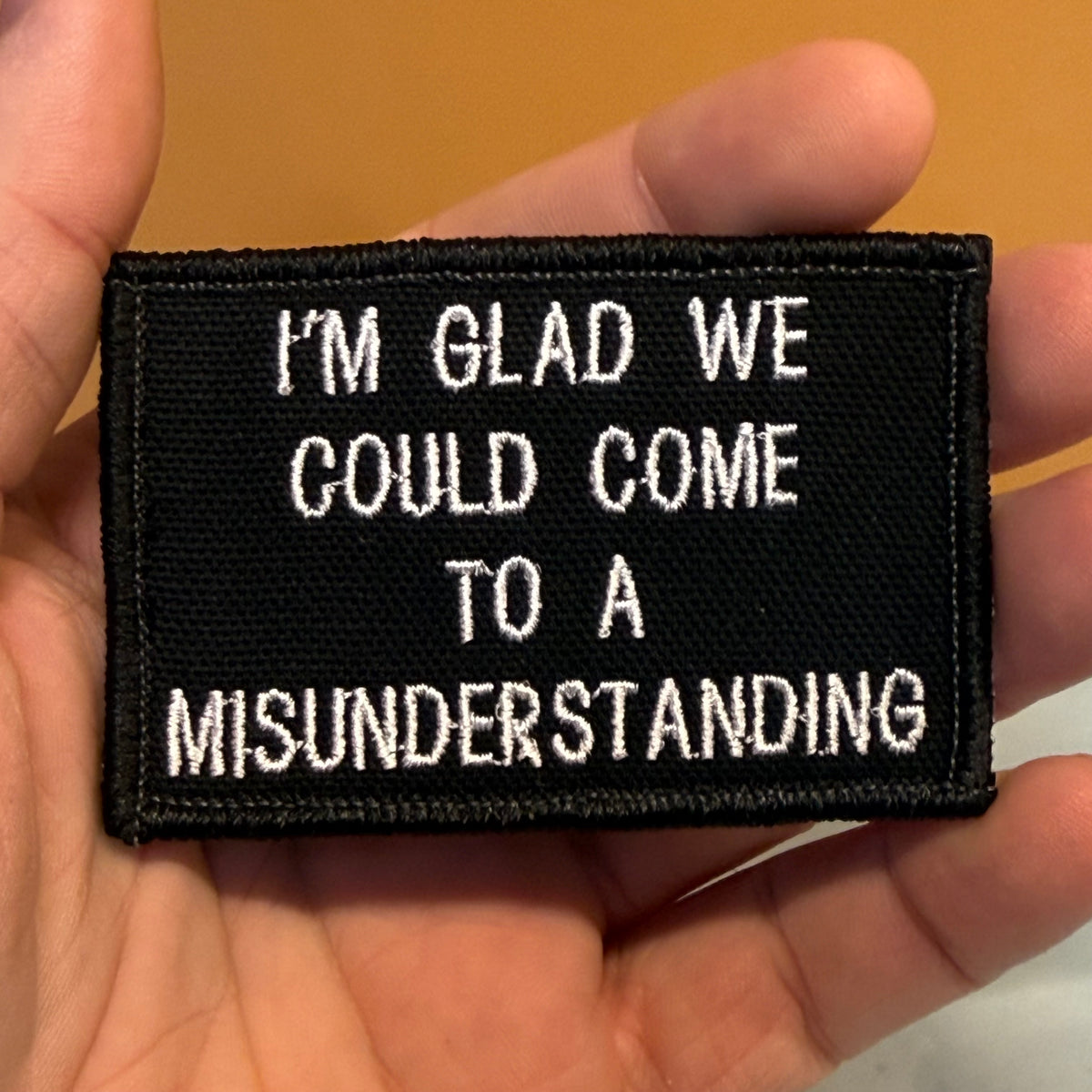 A black rectangular embroidered patch held in a hand, featuring the white text 'I'M GLAD WE COULD COME TO A MISUNDERSTANDING'.