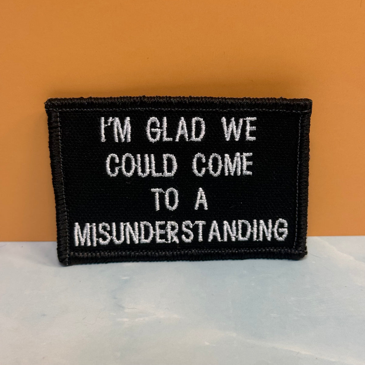 A rectangular black embroidered patch with white text reading 'I'M GLAD WE COULD COME TO A MISUNDERSTANDING', photographed against an orange background and light gray surface.