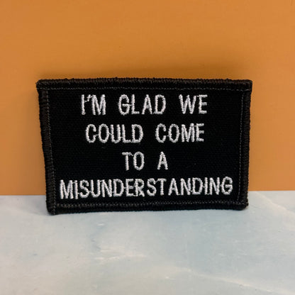 A rectangular black embroidered patch with white text reading 'I'M GLAD WE COULD COME TO A MISUNDERSTANDING', photographed against an orange background and light gray surface.