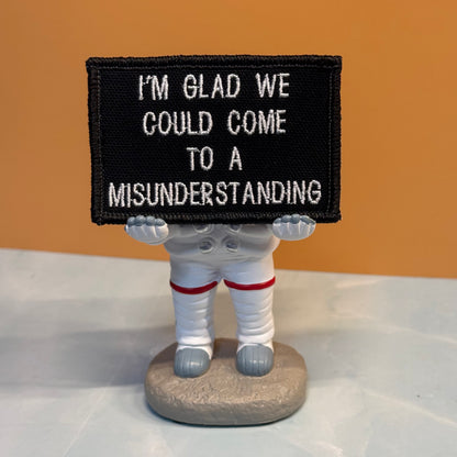 A small astronaut figurine holding a black rectangular patch with white embroidered text that reads, 'I'M GLAD WE COULD COME TO A MISUNDERSTANDING.' The figurine is dressed in a white spacesuit with red stripes and stands on a round gray base against an orange wall and light gray tabletop.