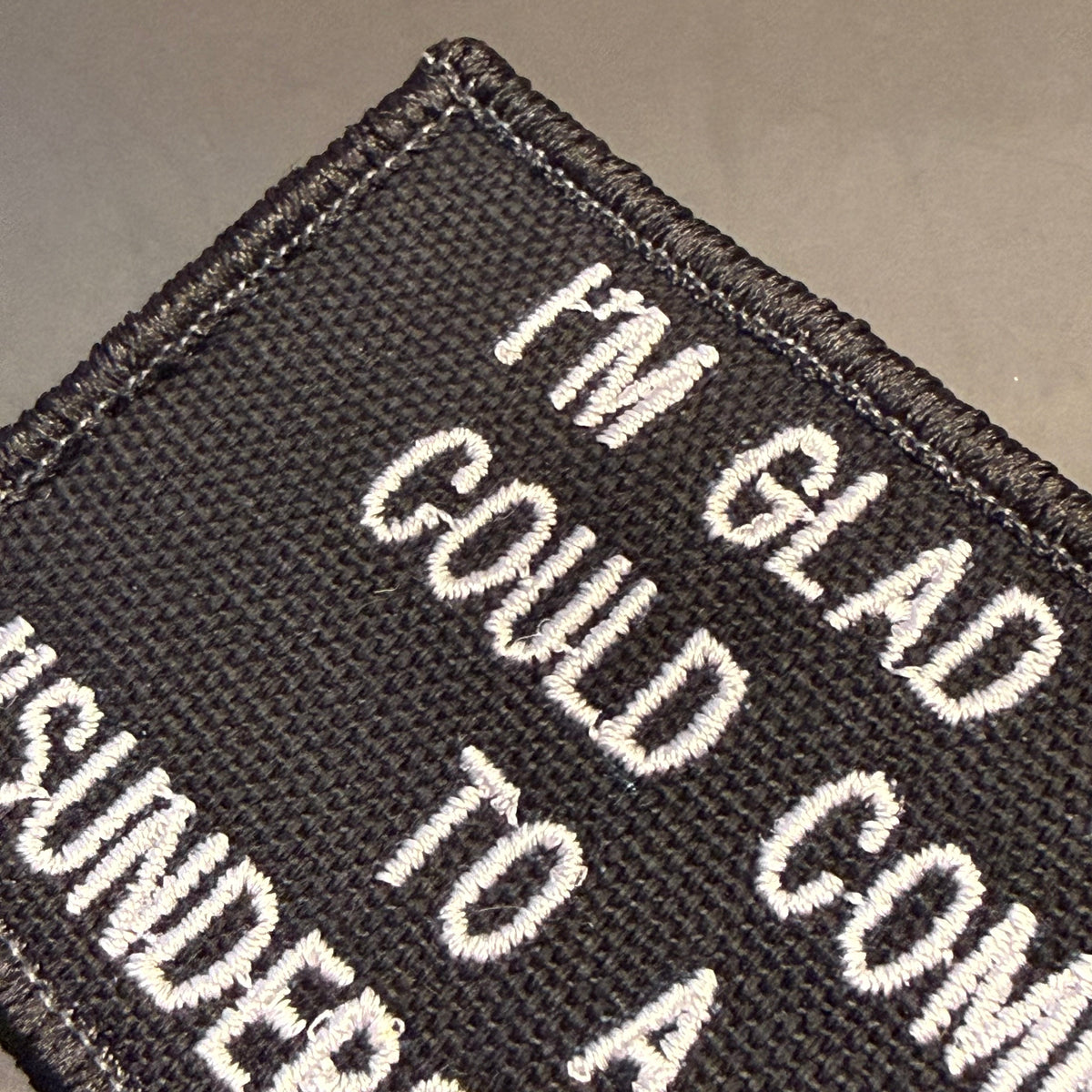 Close-up of a black embroidered patch with white text reading 'I'M GLAD COULD COME TO A SUUNDER' on a neutral gray background.