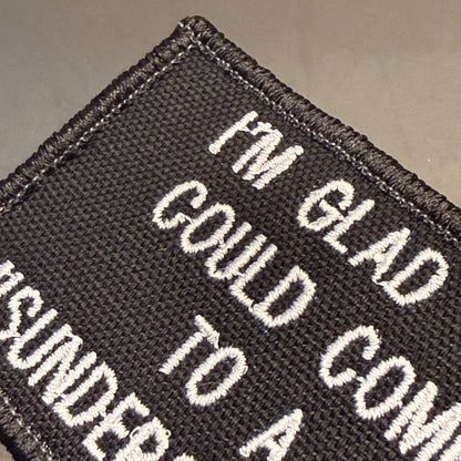 Close-up of a black embroidered patch with white text reading 'I'M GLAD COULD COME TO A SUUNDER' on a neutral gray background.