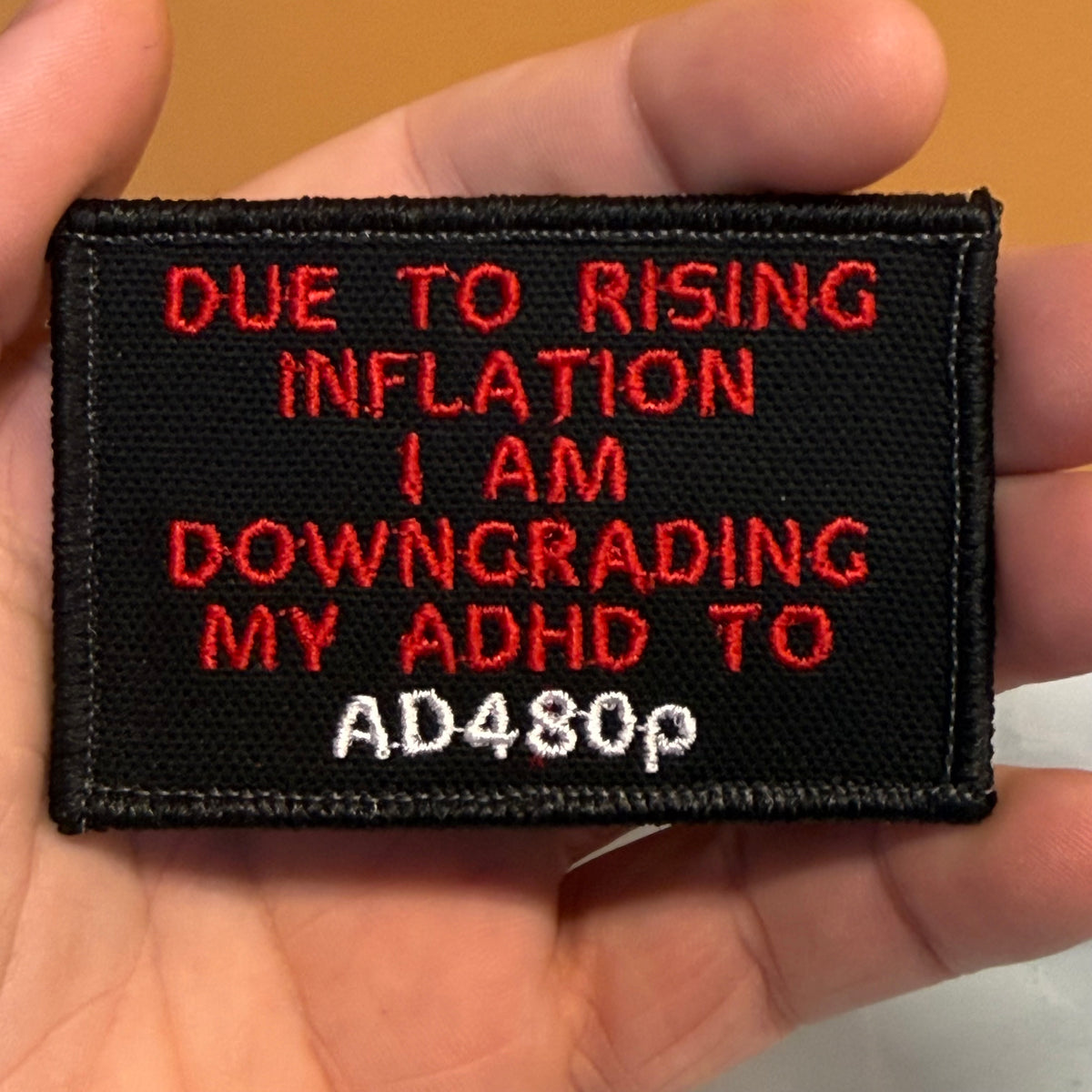 A close-up of a hand holding a rectangular black embroidered patch with red and white text that reads, 'DUE TO RISING INFLATION I AM DOWNGRADING MY ADHD TO AD480p'.