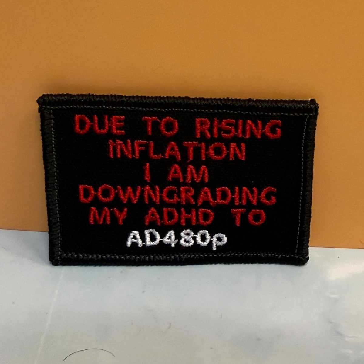 A rectangular black embroidered patch with a stitched border, featuring red and white text that reads, 'DUE TO RISING INFLATION I AM DOWNGRADING MY ADHD TO AD480p' against a two-tone orange and white background.