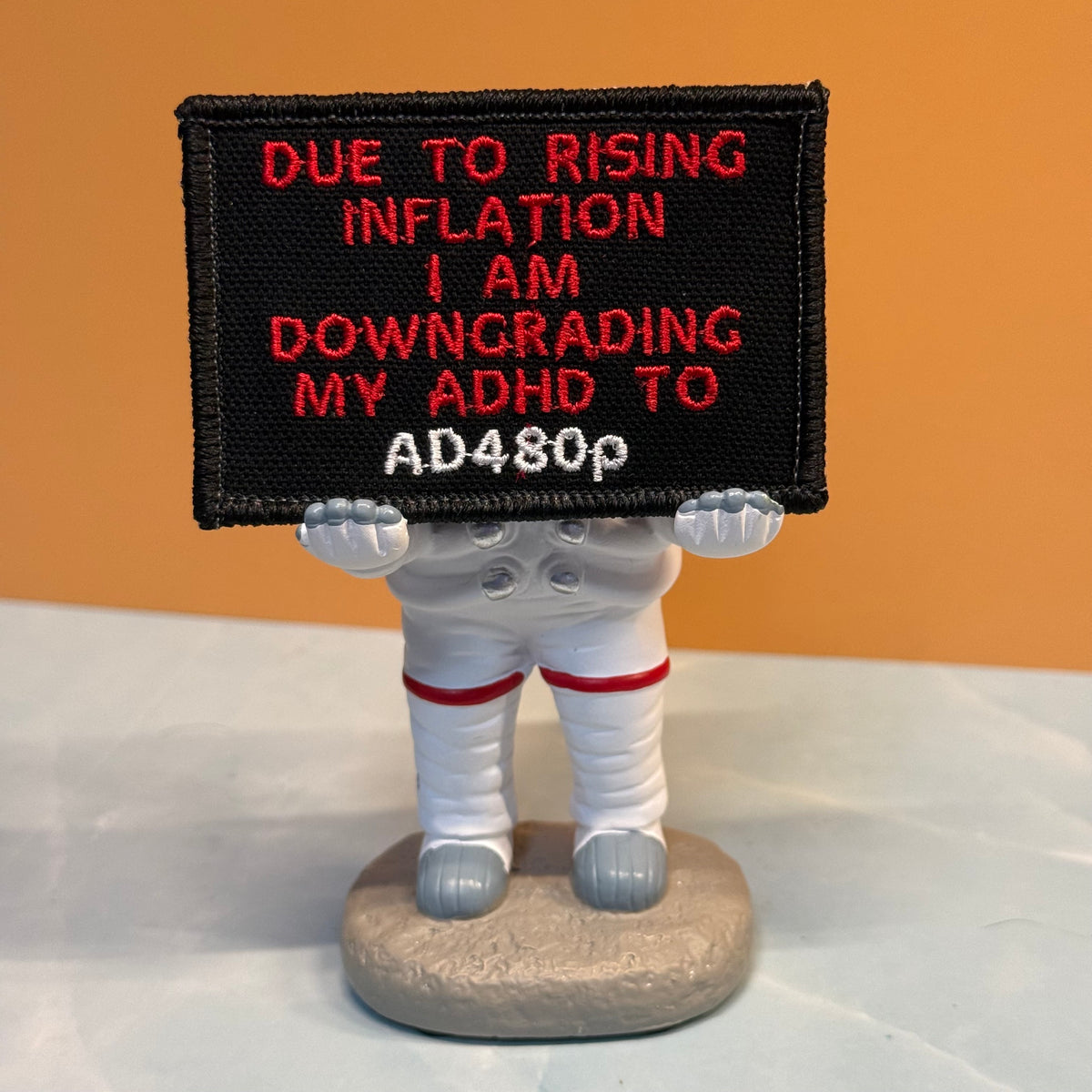 A novelty astronaut figurine holding a black embroidered sign with red and white text that reads, 'DUE TO RISING INFLATION I AM DOWNGRADING MY ADHD TO AD480p.' The figurine stands on a round base against an orange background and white surface.