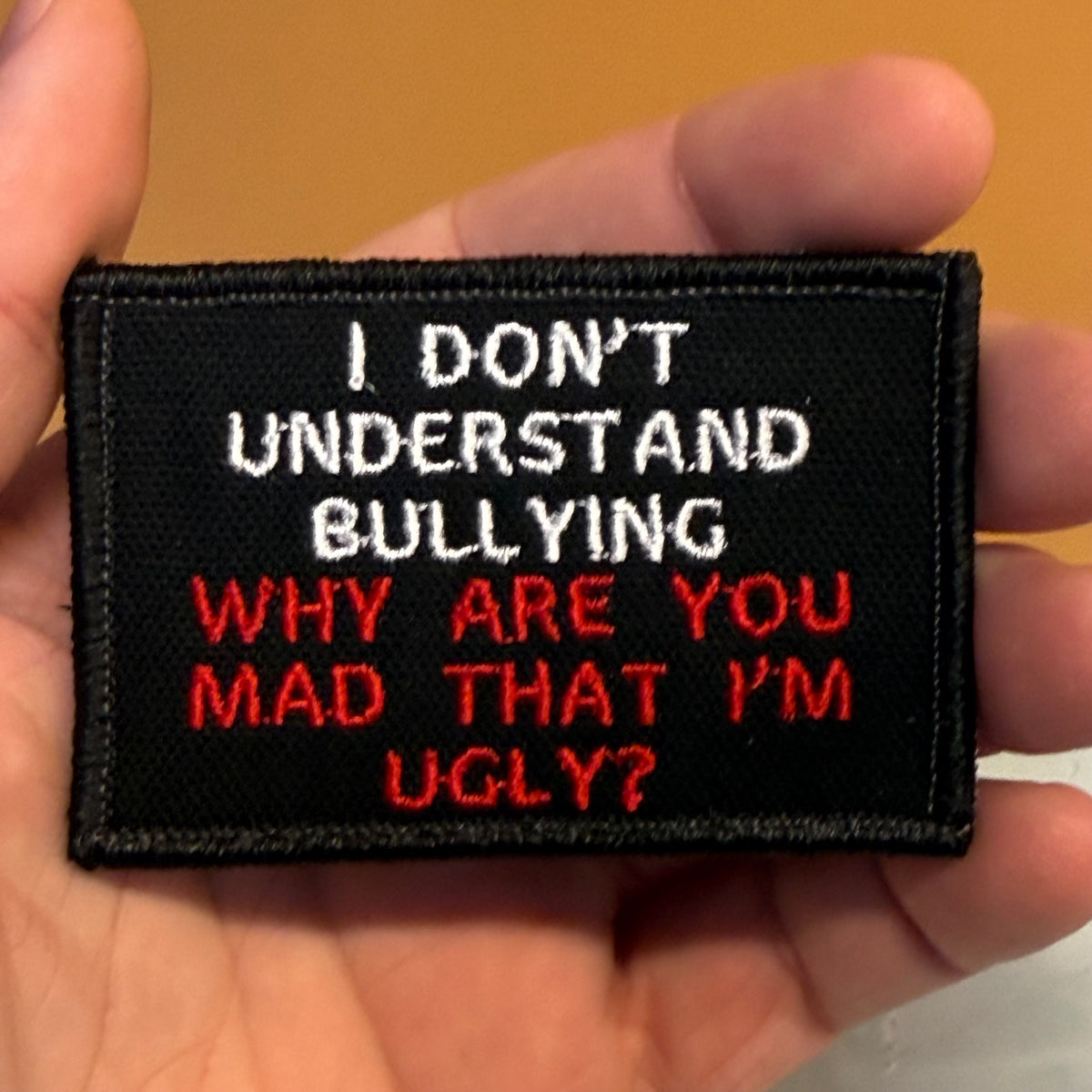 A black embroidered patch held in a hand, featuring the text 'I DON'T UNDERSTAND BULLYING WHY ARE YOU MAD THAT I'M UGLY?' in white and red lettering.