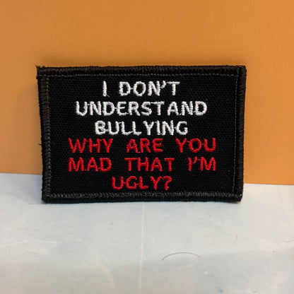 A rectangular black embroidered patch with white and red text reading, 'I DON'T UNDERSTAND BULLYING WHY ARE YOU MAD THAT I'M UGLY?' photographed against an orange background and light gray surface.