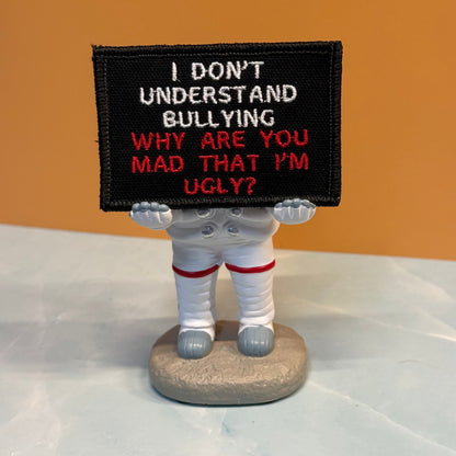 A small figurine of an astronaut holding a black rectangular sign with embroidered text reading, 'I DON'T UNDERSTAND BULLYING WHY ARE YOU MAD THAT I'M UGLY?' The astronaut is white with red stripe accents, standing on a round gray base against an orange background.