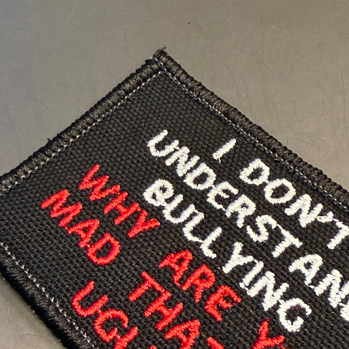 A close-up of a black embroidered patch with white and red text reading 'I DON'T UNDERSTAND WHY MAD ARE THA Y' on a dark gray background.