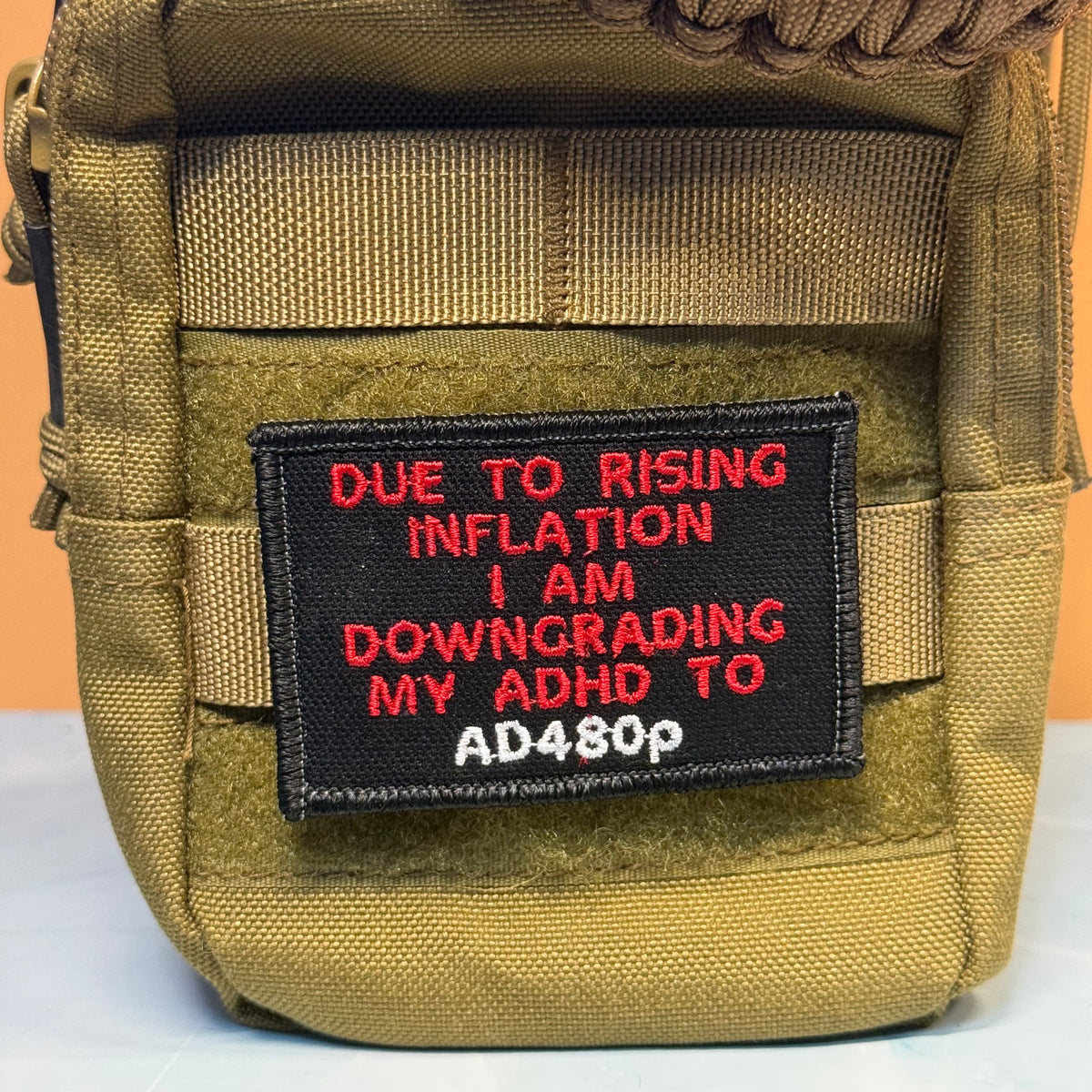 A close-up of a tan tactical pouch featuring a black rectangular patch with red and white embroidered text that reads, 'DUE TO RISING INFLATION I AM DOWNGRADING MY ADHD TO AD480p'.