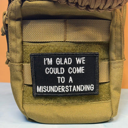A close-up of a tan tactical pouch with a black rectangular patch that reads, 'I'M GLAD WE COULD COME TO A MISUNDERSTANDING' in white text.