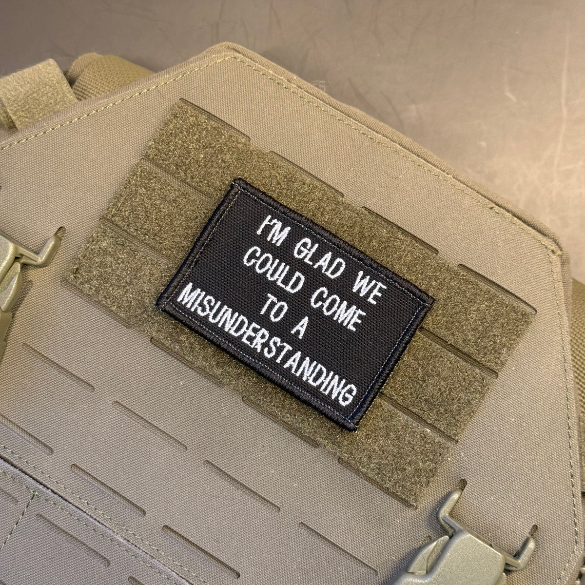 A close-up of a tactical vest or bag in coyote tan with a black rectangular embroidered patch that reads, 'I'M GLAD WE COULD COME TO A MISUNDERSTANDING.'.
