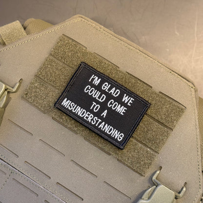 A close-up of a tactical vest or bag in coyote tan with a black rectangular embroidered patch that reads, 'I'M GLAD WE COULD COME TO A MISUNDERSTANDING.'.