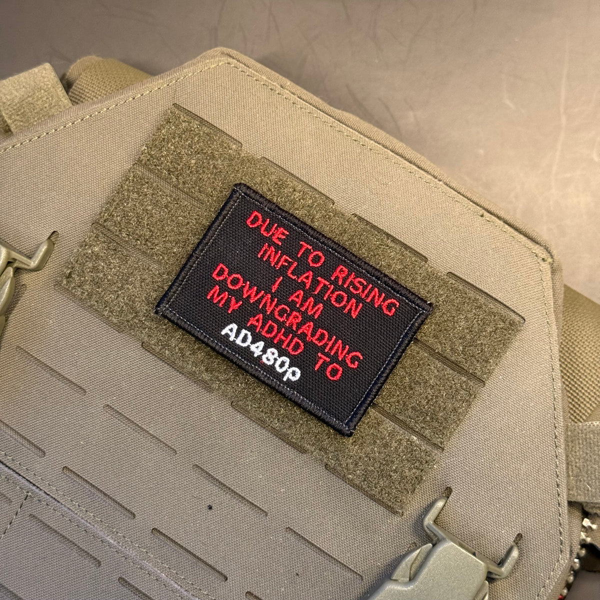 A close-up of a tactical bag in tan canvas with a black rectangular patch sewn onto MOLLE webbing. The patch features red embroidered text that reads 'DUE TO RISING INFLATION I AM DOWNGRADING MY ADHD TO AD480p'.