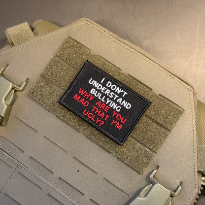 A rectangular black embroidered patch with the text 'I DON'T UNDERSTAND BULLYING WHY ARE YOU MAD THAT I'M UGLY?' in white and red lettering, attached to tan tactical webbing on a beige bag or vest.