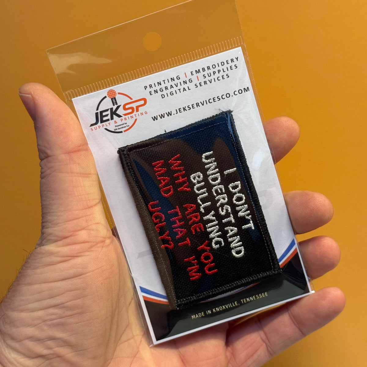 A hand holds a packaged embroidered patch against a solid orange background. The patch reads “I DON’T UNDERSTAND WHY YOU ARE YOU MAD UGLY” in white and red on a black background, and the packaging shows the brand JEKSP.