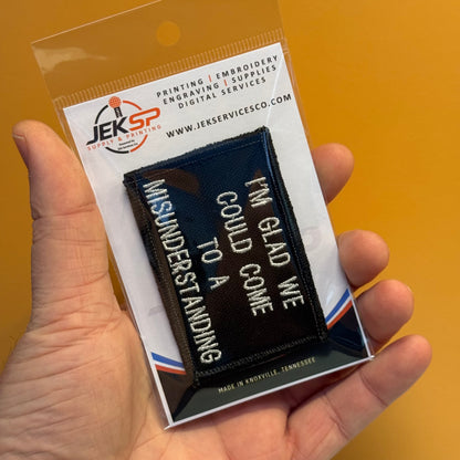 A hand holds a clear plastic blister pack containing a black embroidered patch with white text that reads, 'I'M GLOD WE COULD COME TO A UNDERSTANDING.' The backing card shows the JEKSP brand and service offerings, against a solid mustard-yellow background.