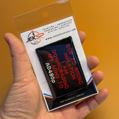 A hand holds a clear plastic blister pack containing a rectangular embroidered patch. The patch has a black background with red and white text reading, 'DUE TO RISING INFLATION I AM A ADDICT TO DOWNING MY ADDICTION TO AD4809.