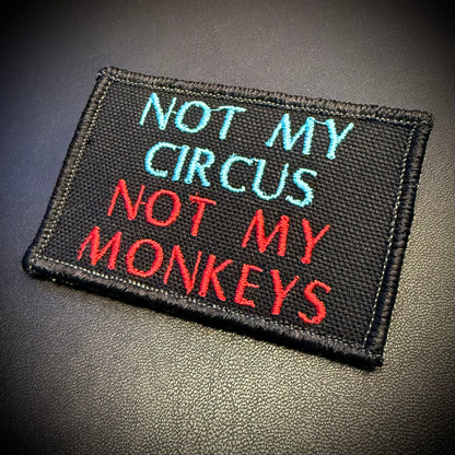 A rectangular black embroidered patch with the text 'NOT MY CIRCUS NOT MY MONKEYS' in light blue and red lettering, shown on a dark textured surface.