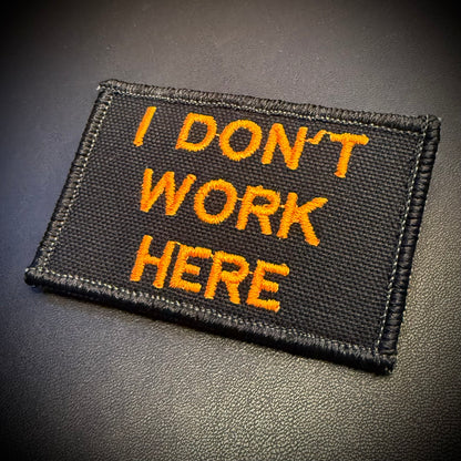 A rectangular black embroidered patch with the text 'I DON'T WORK HERE' in bright orange lettering, shown on a dark textured surface.