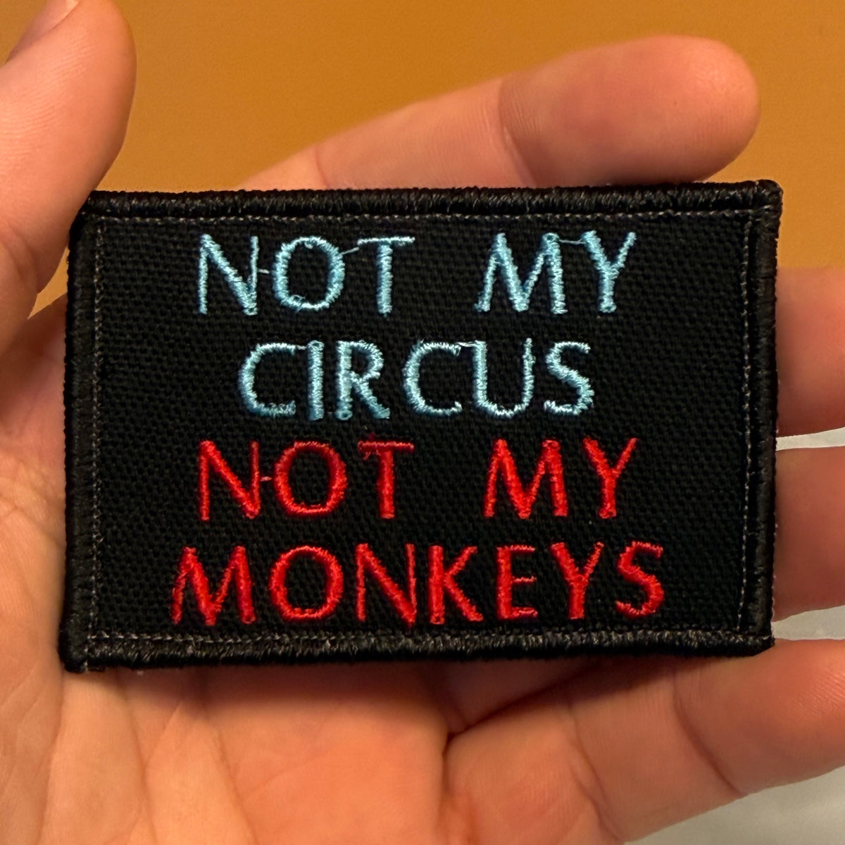 A rectangular black embroidered patch held in a hand, featuring the text 'NOT MY CIRCUS NOT MY MONKEYS' in light blue and red lettering.