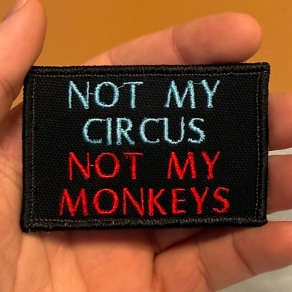 A rectangular black embroidered patch held in a hand, featuring the text 'NOT MY CIRCUS NOT MY MONKEYS' in light blue and red lettering.
