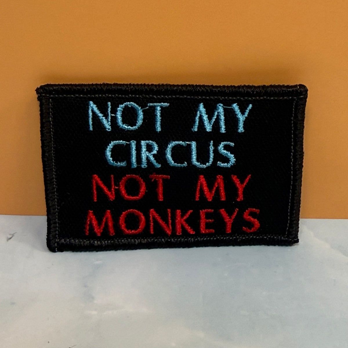 A rectangular black embroidered patch with the text 'NOT MY CIRCUS NOT MY MONKEYS' in light blue and red thread, displayed on a white marble-like surface against an orange background.